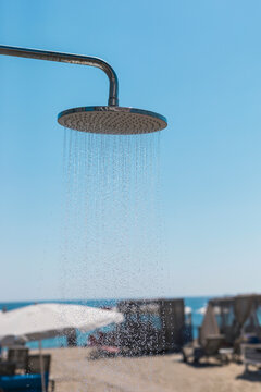 Fototapeta A metal shower head dispenses water in a beach setting with a bright blue sky above. Sunbeds and umbrellas are visible near the shore, inviting relaxation and refreshment