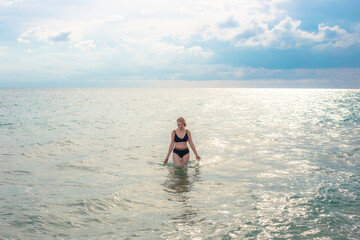 A woman in a bikini stands in the ocean admiring the water surrounded by soft waves under a cloudy...