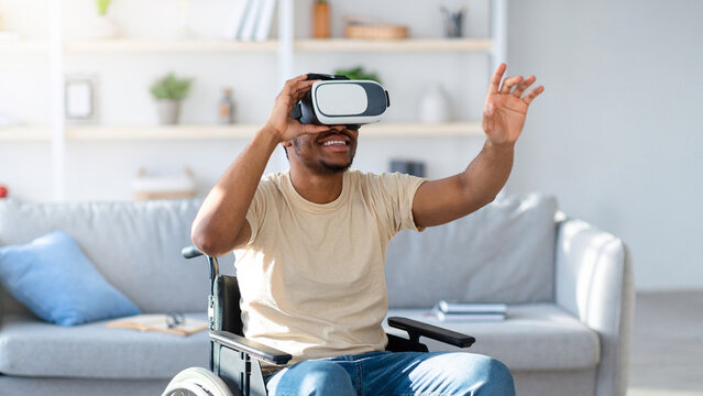 Young black man in wheelchair exploring augmented reality in VR goggles at home. Paraplegic African American guy playing virtual game, using modern technologies for fun
