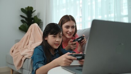 Happy girl playing game and sitting while mom lie on sofa by using laptop screen. Caucasian parent...