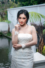 A beautiful Indonesian bride in a white wedding dress adorned with lace and pearl details, posing...