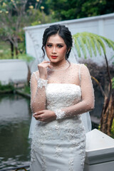 An Indonesian bride wearing a white lace wedding dress with pearl details, striking a graceful and...