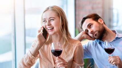 Young woman speaking on mobile phone during dull date, her boyfriend feeling lonely and bored at coffee shop. Young couple having communication problems on romantic dinner at restaurant
