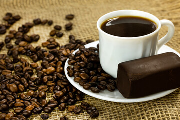 White cup of black coffee on a white saucer with coffee beans and a rectangular chocolate bar on a burlap surface.
