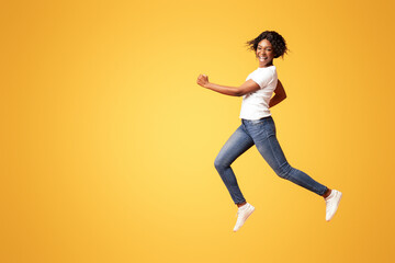 Active black lady in casual outfit running over orange studio background, full length shot. Cheerful african american young woman in jeans and white t-shirt having fun on her own