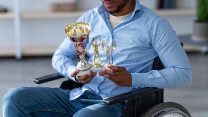 Cropped view of black guy in wheelchair holding different sports trophies, feeling depressed over...