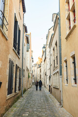 Two people stroll down a narrow, cobblestone-paved lane in Tours. Ancient buildings with closed shutters stand on either side. It is an overcast day in early spring