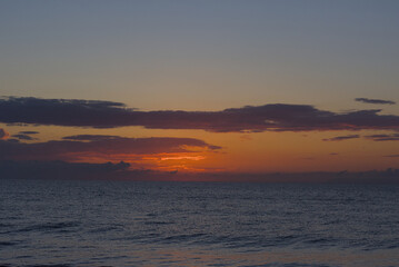 Obraz premium The sun rises on the horizon over a calm, dark ocean, painting the sky with warm orange, pink, and purple hues and scattering light across the water.