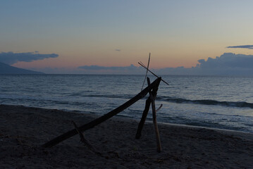 A makeshift structure of driftwood stands on a sandy beach at dusk, with gentle waves rolling in and mountains visible in the distance under a cloudy sky.