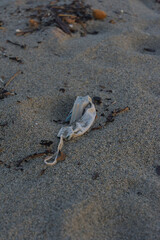 A dirty, crumpled white disposable face mask lies on a sandy beach, surrounded by small pieces of dark debris.