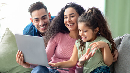 Happy Arabic Family Of Three Making Video Call With Laptop At Home, Joyful Middle Eastern Parents And Their Little Daughter Having Web Chat With Family, Enjoying Online Communication, Closeup