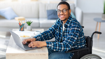 Cheerful black man in wheelchair using laptop for online job from home, having business meeting on web. African American with disability freelancer working on remote project