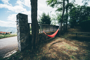 Bright red hammock hanging between trees in a shaded corner of a small park, near a stone fence...