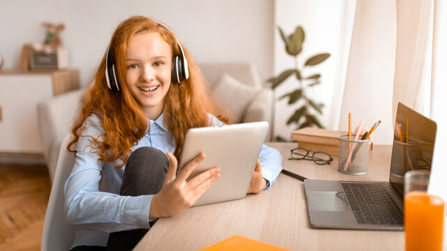 Portrait Of Smiling Red-Haired Teenage Girl Sitting At Desk At Home, Holding And Using Digital Tablet, Listening To Music Or Podcast In Wireless Headphones, Looking At Camera, Relaxing And Resting