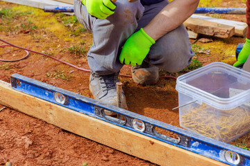 Construction worker adjusts wood beam using level spirit tool on dirt surface with construction materials nearby.