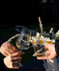 Celebration moment, clinking wine glasses, hands toasting, flowers in vase, romantic evening, outdoor dinner, natural light, festive mood, garden party, white wine, birthday, event, spring, summer