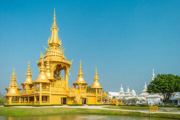 Naklejka premium Golden Ganesha temple in Wat Rong Khun complex with ornate spires and traditional architecture surrounded by garden and pond under blue sky in sunny day, Chiang Rai, Thailand. White temple complex