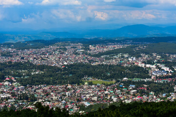 Shillong city nestled amidst hills and lush greenery, showcasing a panoramic view under a cloudy sky.