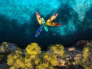 Aerial drone view of colorful kayaks with people paddling on crystal clear turquoise sea near rocky coast and trees. Summer outdoor adventure in Mallorca, Balearic Islands, Spain. Top view of canoes