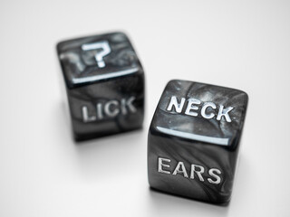 Two black marble dice with embossed words suggesting intimate actions and body parts on a white...