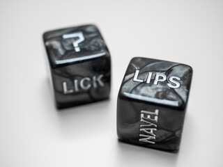 Two black marble dice with embossed words suggesting intimate actions and body parts on a white...
