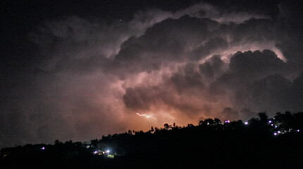 Vivid lightning striking through dark night sky over silhouetted hills with distant lights, capturing nature's power.