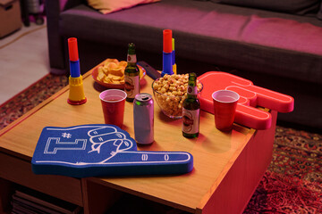 Closeup showing table with snacks, popcorn, potato chips, beer bottles, soda can, plastic cups, colorful foam hands and vuvuzelas, suggesting sports viewing or party gathering
