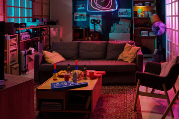 Modern living room featuring empty gray sofa and armchair, illuminated by neon lights, coffee table holding snacks, drinks, party cups, and tabletop game, shelves with books and guitars in background