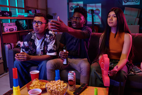 Young adult Caucasian man, young adult Black man, and young adult Asian woman sitting on sofa playing video games together, holding controllers and snacks visible on table