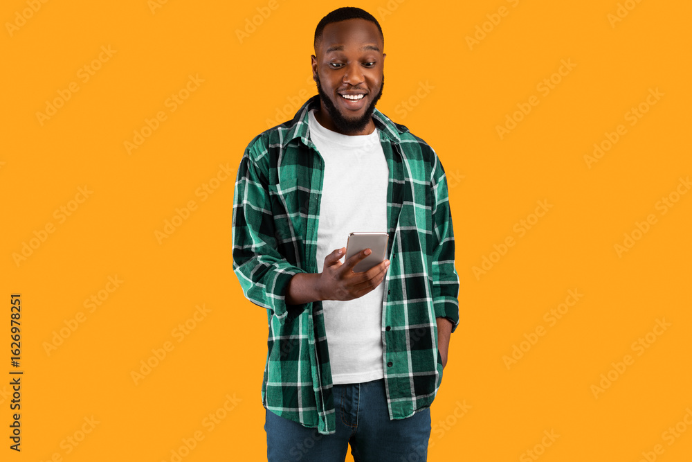 Aufkleber cheerful black man using smartphone browsing internet and texting standing over yellow studio backgr