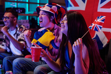 Group of young adult multiethnic friends cheering and celebrating while watching sports event indoors, smiling and holding British flags, showing excitement and unity