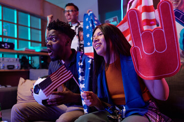 Young Black man holding soccer ball and small American flag sitting next to young Asian woman cheering with foam hand while watching sports event, excited middle aged Caucasian man standing behind