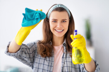 Portrait of smiling young woman in rubber gloves cleaning glass with spray detergent, indoors....