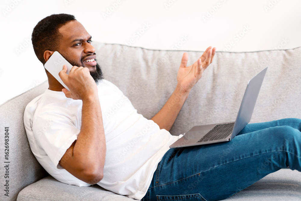Poster side view of discontented african guy talking on cellphone using laptop and having problem with inte