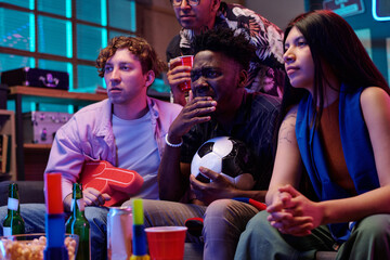 Multiethnic group of young adults watching sports game on television, Black man holding soccer ball...