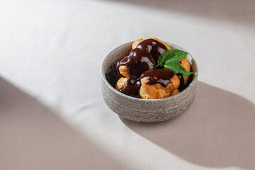 Golden profiteroles generously drizzled with chocolate sauce and decorated with mint leaves, served in a stone-textured bowl on a light background