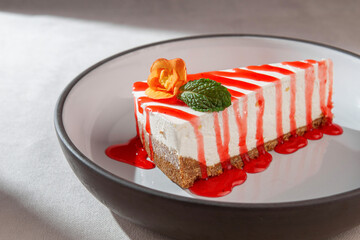 Cheesecake slice with graham crust, strawberry syrup, mint leaf and edible flower served on a modern ceramic plate in soft natural light