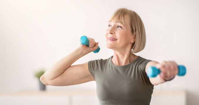 Domestic sports training. Positive senior woman doing exercises with dumbbells, strengthening her body at home. Fit mature lady working on her biceps muscles, staying healthy during covid quarantine