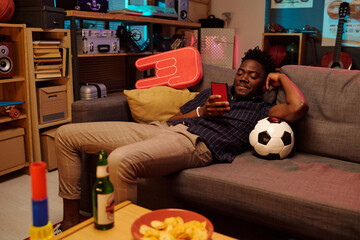 Young adult Black man reclining on sofa holding smartphone and resting head on soccer ball,...