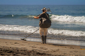 A man using a metal detector on a beach with the ocean and waves in the background