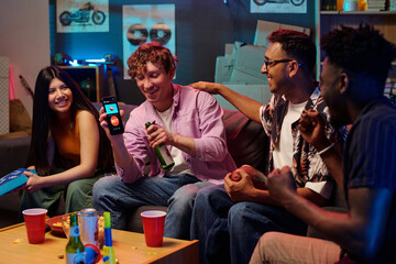Group of young adult men and woman sitting on couch playing smartphone game together, smiling and laughing, holding drinks and snacks, multiethnic friends enjoying leisure time indoors