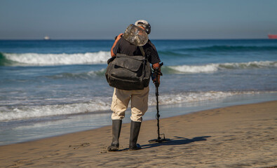 A man using a metal detector on a beach with the ocean and waves in the background