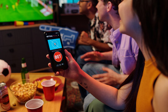 Young adult Asian woman holding smartphone with sports betting app while sitting with diverse group of young adults watching soccer match on television, snacks and drinks on table