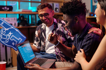 Group of young adults including Black man, Caucasian man, and Asian woman celebrating while watching sports betting results on laptop, smiling and interacting in casual setting