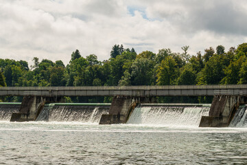 Water flows over a dam in a lush green landscape under a cloudy sky during the day