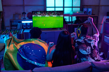 Three young adults including Black man, Asian woman, and Caucasian man sitting on couch watching soccer match on television, cheering and holding Brazilian flag indoors