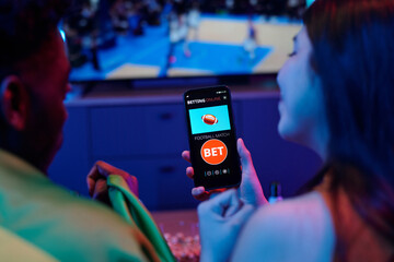 Young adult Black man and young adult Caucasian woman watching sports event on television while woman holding smartphone displaying online football betting application interface