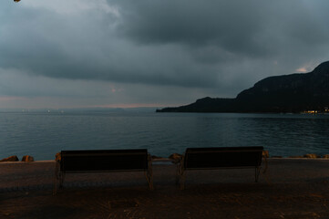 A moody, atmospheric scene of two empty benches placed at the edge of Lake Garda, Italy, captured...