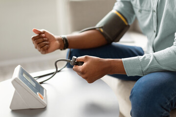 Unrecognizable Black Man Measuring Arterial Blood Pressure Having Hypertension Health Issue Sitting On Couch Indoors. Hypotension, Low And High Blood-Pressure Problem Concept. Cropped