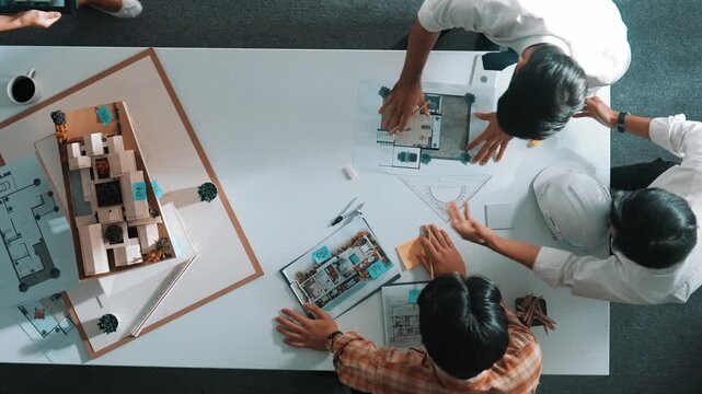 Group of skilled designer looking at model while discussing about building. Top view of architect engineer explain idea while talking and writing blue print scattering at meeting table. Alimentation.